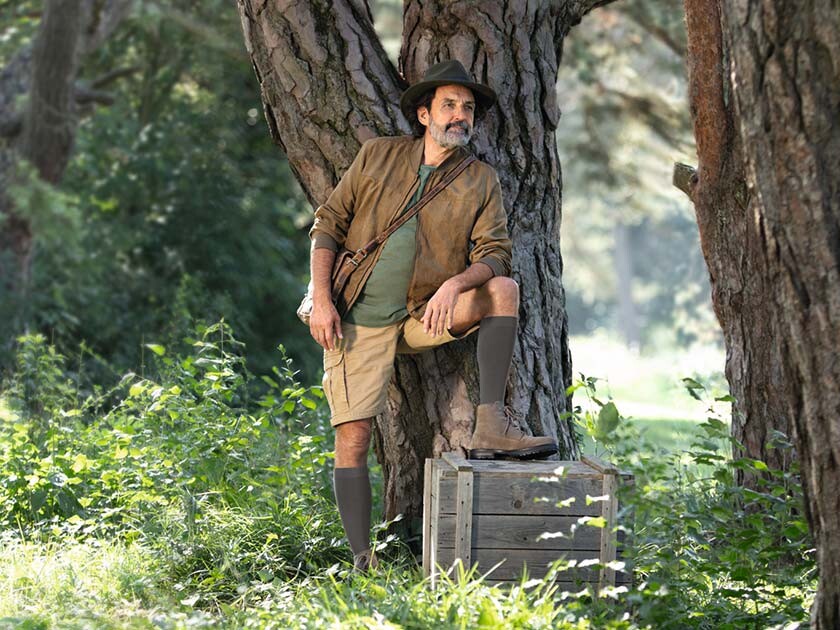 Ein Mann mit Entdecker-Outfit steht in der Natur und schaut in die Ferne. Er trägt kniehohe Kompressionsstrümpfe.