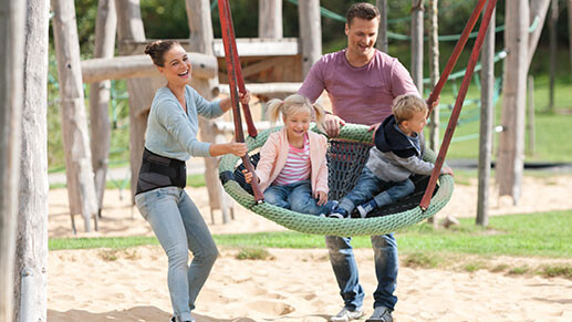 Eine Familie auf dem Spielplatz. Die Kinder schaukeln. Die Mutter trägt eine Rückenbandage