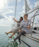 Two people are sitting relaxed at the edge of a sailing boat, looking out over the water; one is wearing compression garments on their arms and legs.