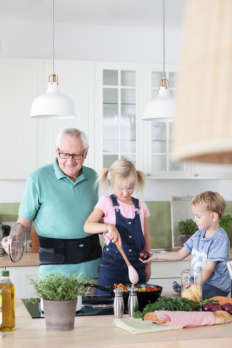 Grandpa is cooking with his grandchildren. He is wearing a back orthosis.